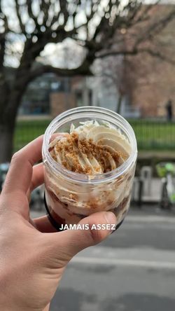 Dessert en pot avec crème chantilly et crumble tenu à la main devant un paysage urbain avec arbres.