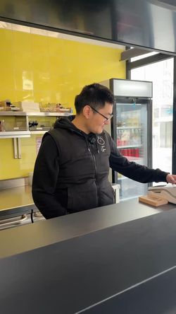 Employé en tenue noire prenant une commande au comptoir dans un restaurant japonais. Fond jaune et vitrine réfrigérée.