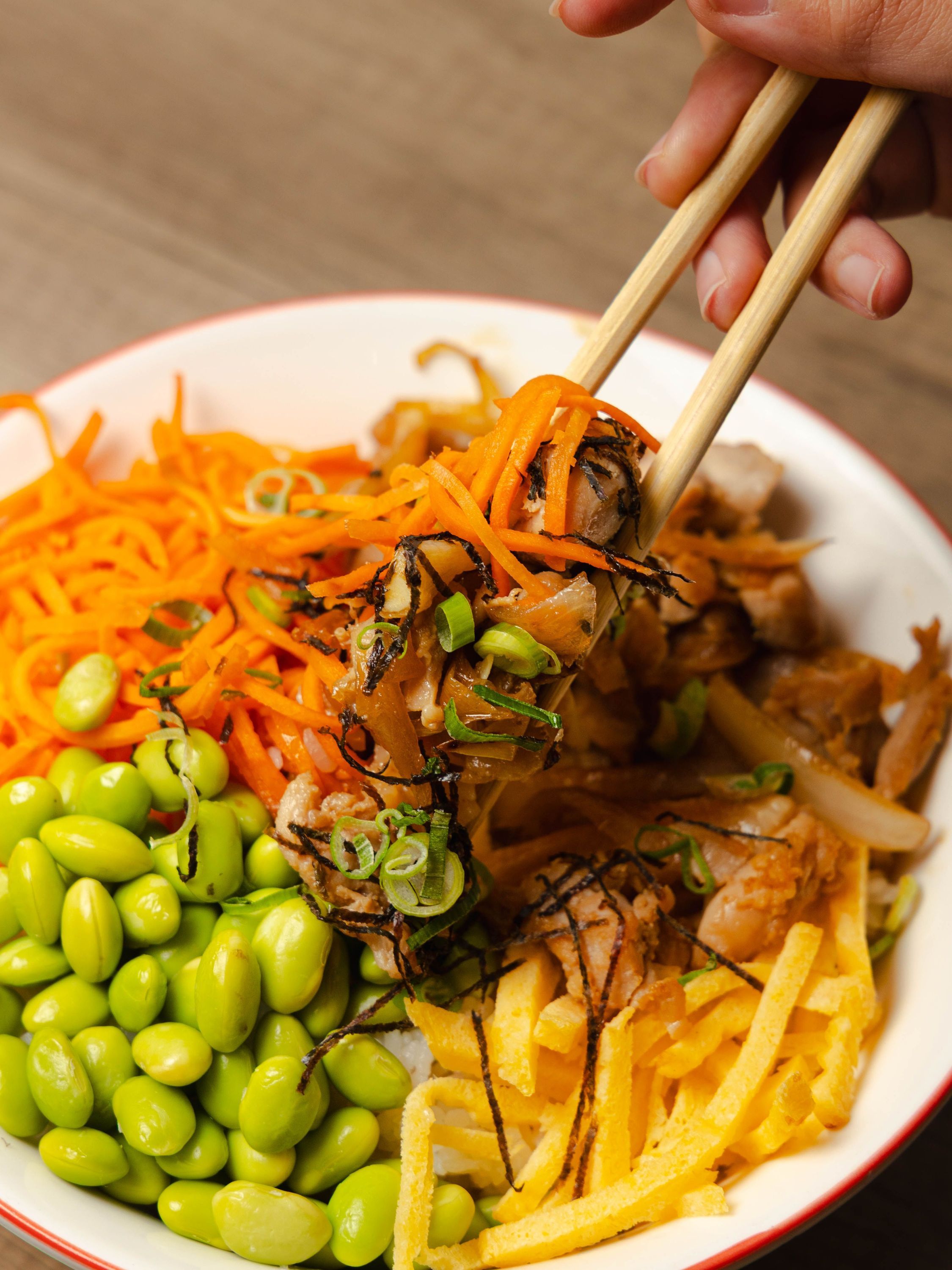 Bol asiatique avec edamame, carottes râpées, oignons verts, et morceaux de poulet, dégusté avec des baguettes.