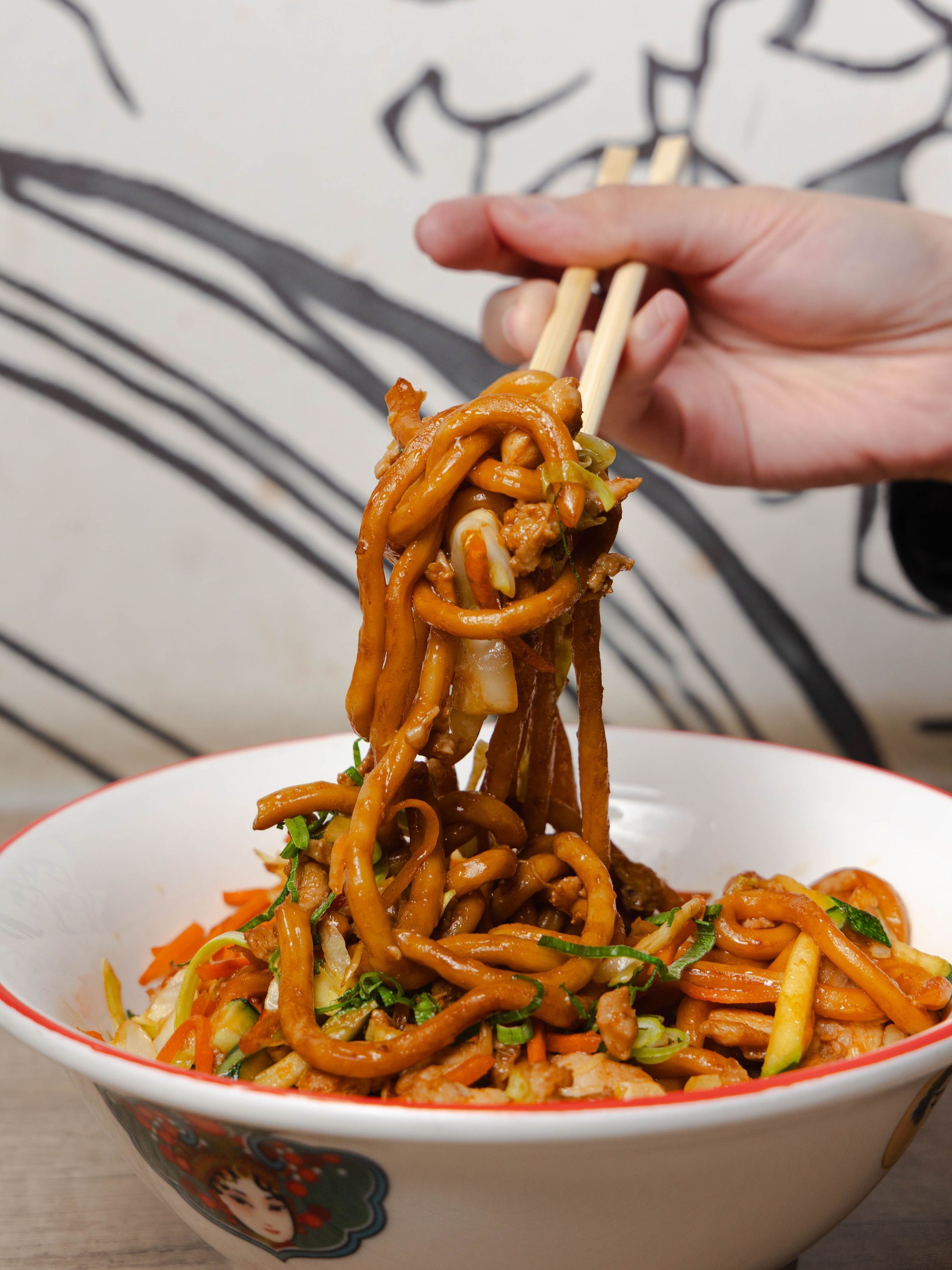 Plat de nouilles sautées servi dans un bol typique japonais, accompagné de légumes variés, avec utilisation de baguettes.