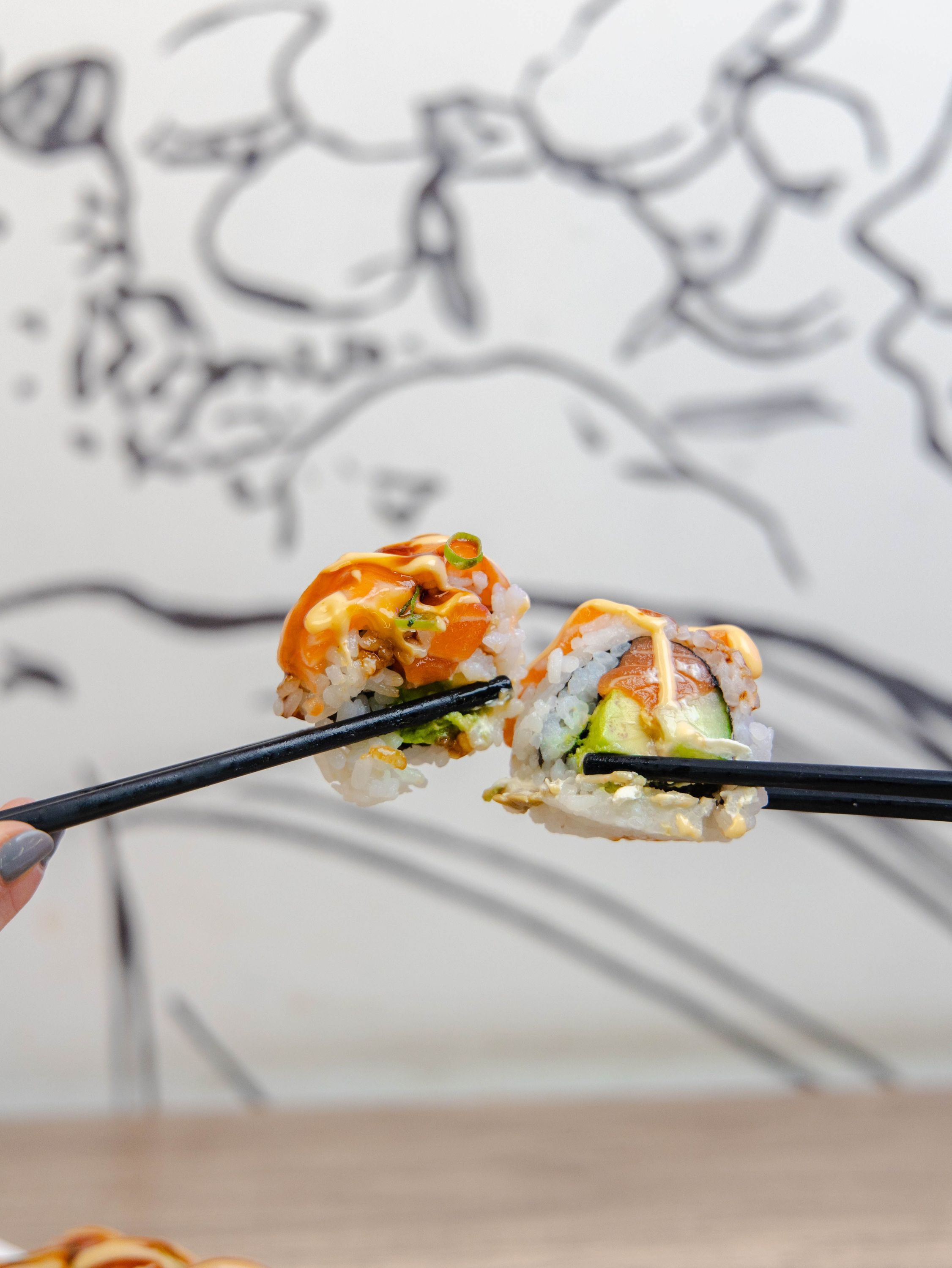 Deux pièces de california rolls tenues par des baguettes, avec une sauce orange visible dessus.