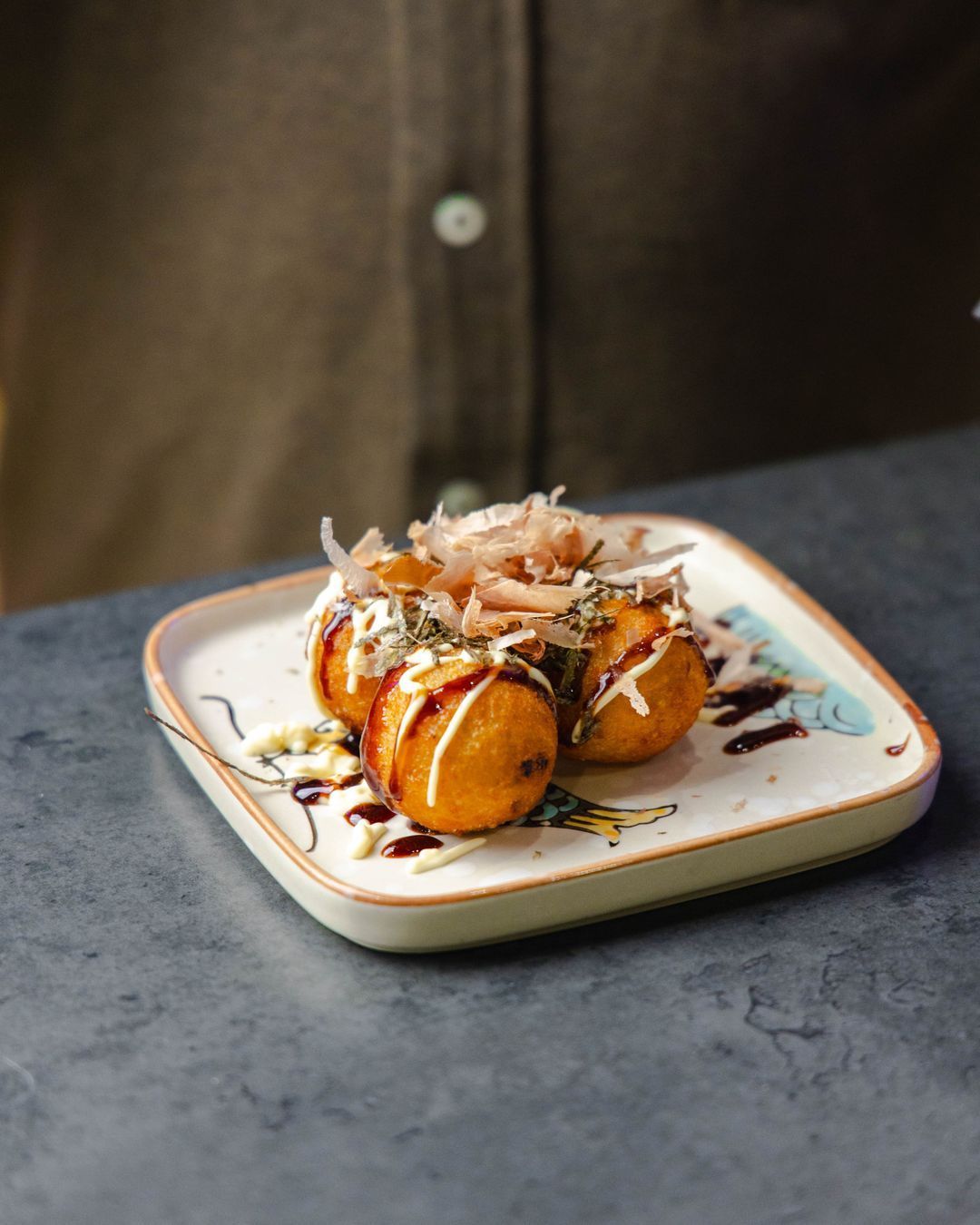 Boulettes japonaises garnies de bonito sur une assiette décorative, avec sauce à l'extérieur.