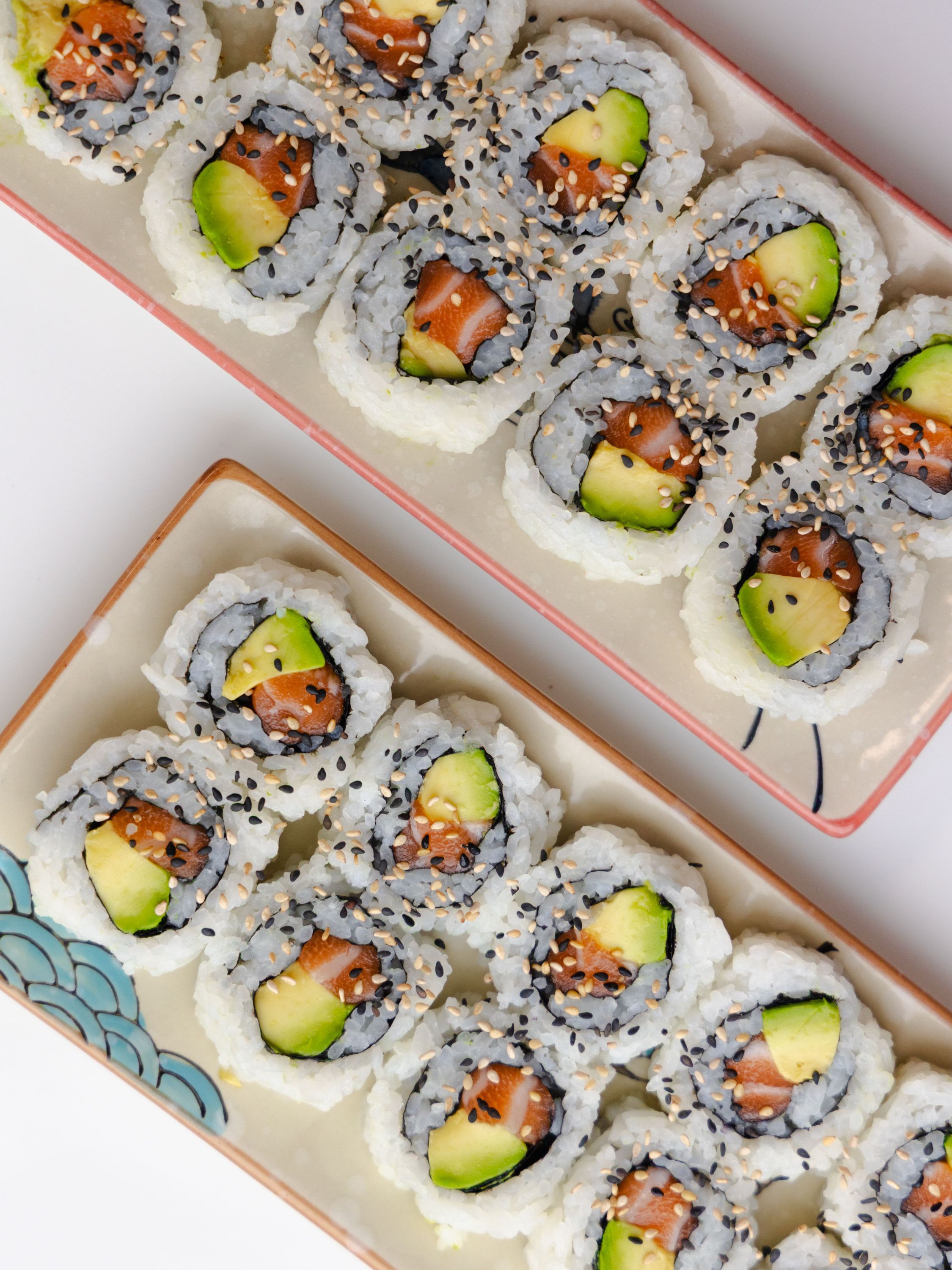 Sushis maki avec avocat et saumon, garnis de graines de sésame, servis sur deux assiettes rectangulaires.