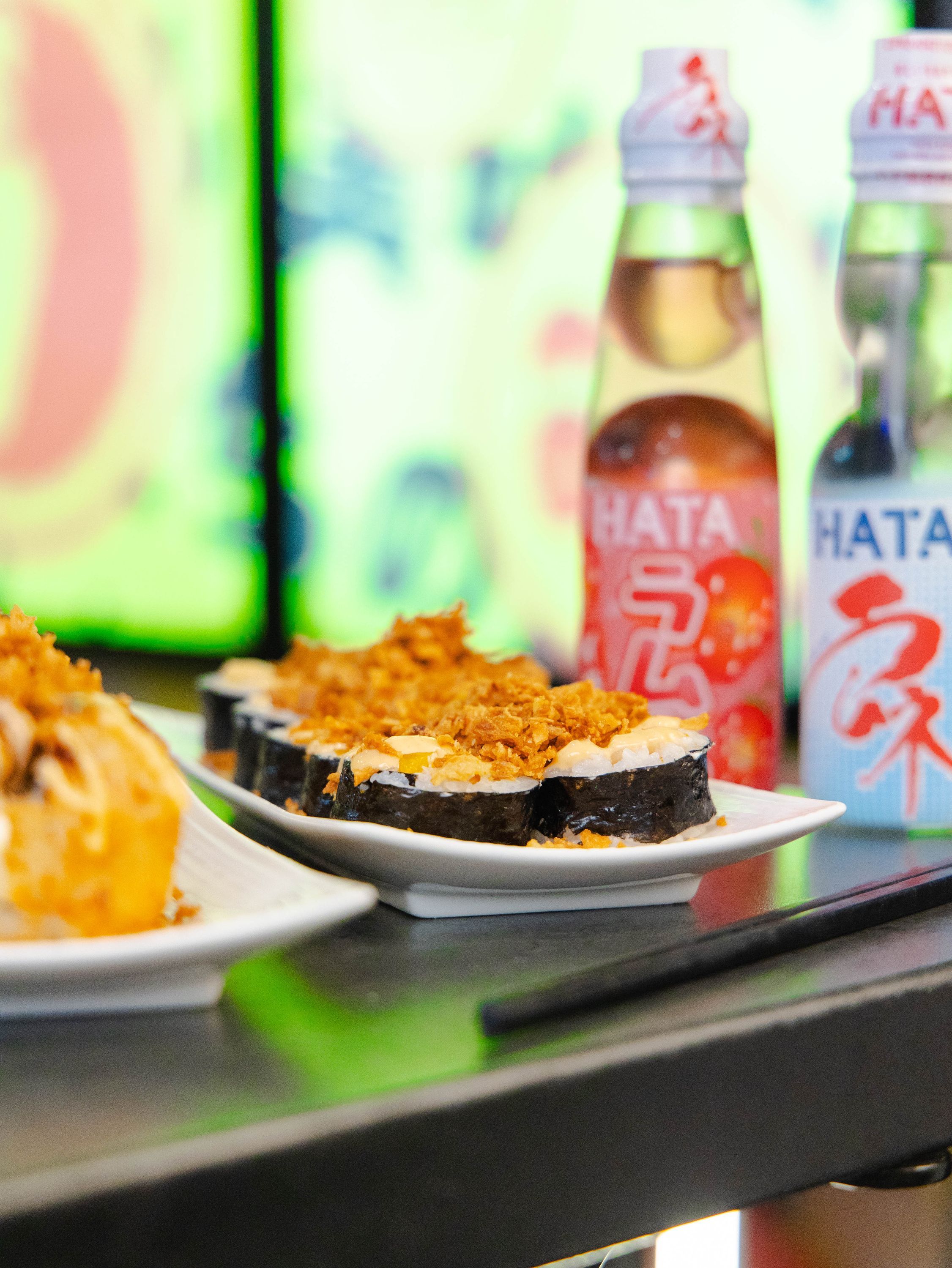 Sushi sur assiettes, en avant-plan, avec des bouteilles de boisson HATA et un fond coloré.