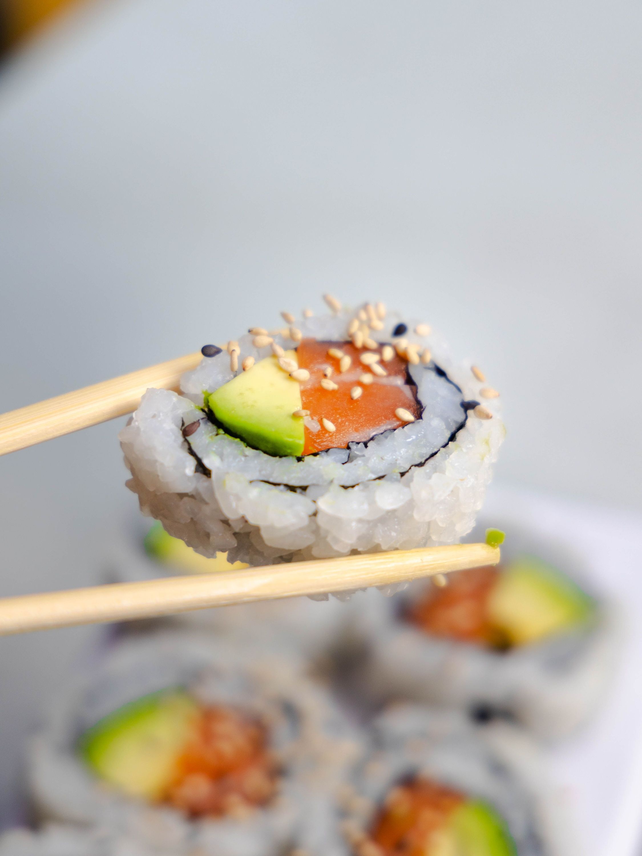 Rondelle de sushi californien avec avocat et saumon tenue par des baguettes, fond flou avec d'autres sushis similaires.