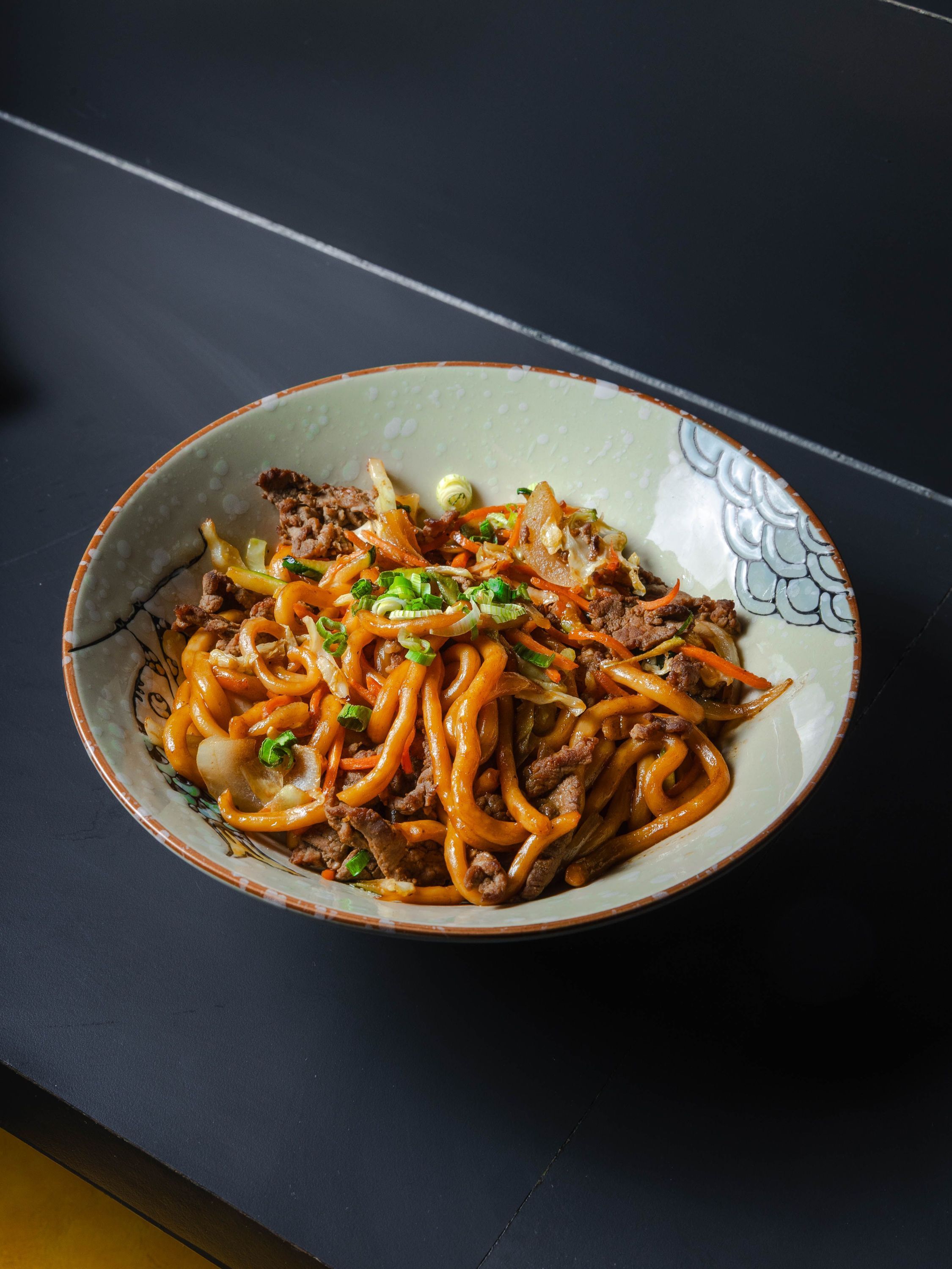Plat asiatique avec nouilles sautées, morceaux de viande et légumes verts dans un bol décoré.