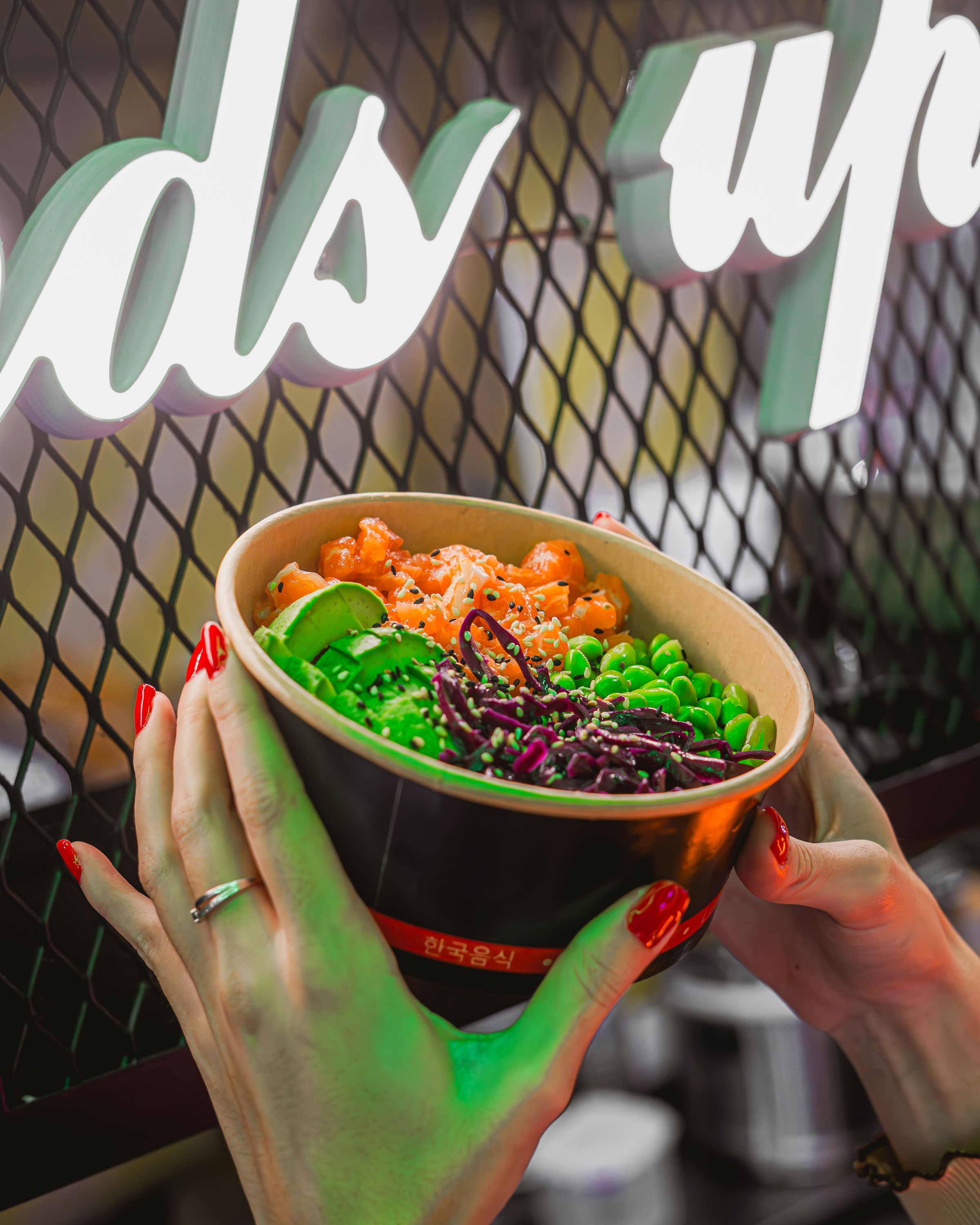 Bol rempli de crevettes, edamame et légumes variés, tenu devant une enseigne lumineuse au restaurant.