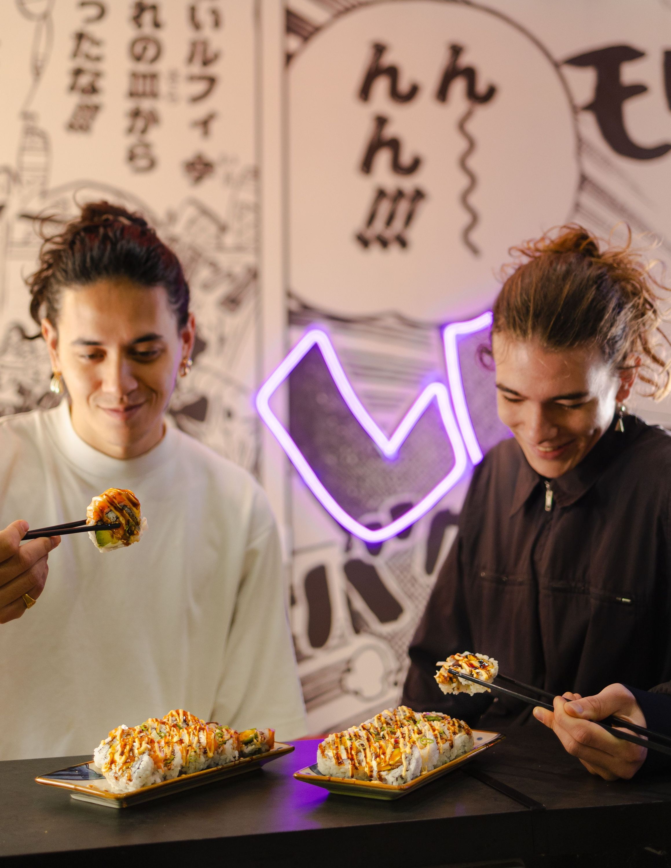 Deux personnes dégustent des California rolls dans un restaurant japonais avec décor mural manga et lumière néon violet.